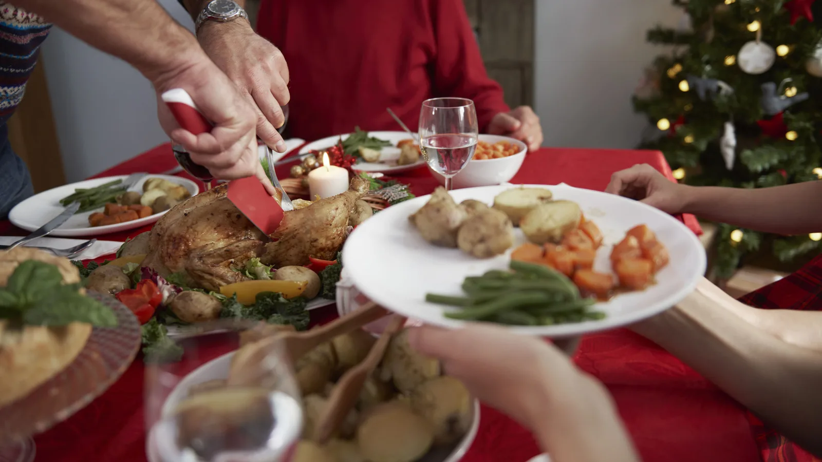 Família à mesa de fim de ano com frango assado sendo fatiado e árvore de Natal ao fundo